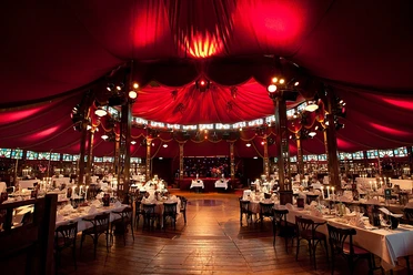 Ein festlich beleuchtetes, luxuriöses Eventzelt mit rot-golden dekoriertem Interieur und gedeckten Tischen.