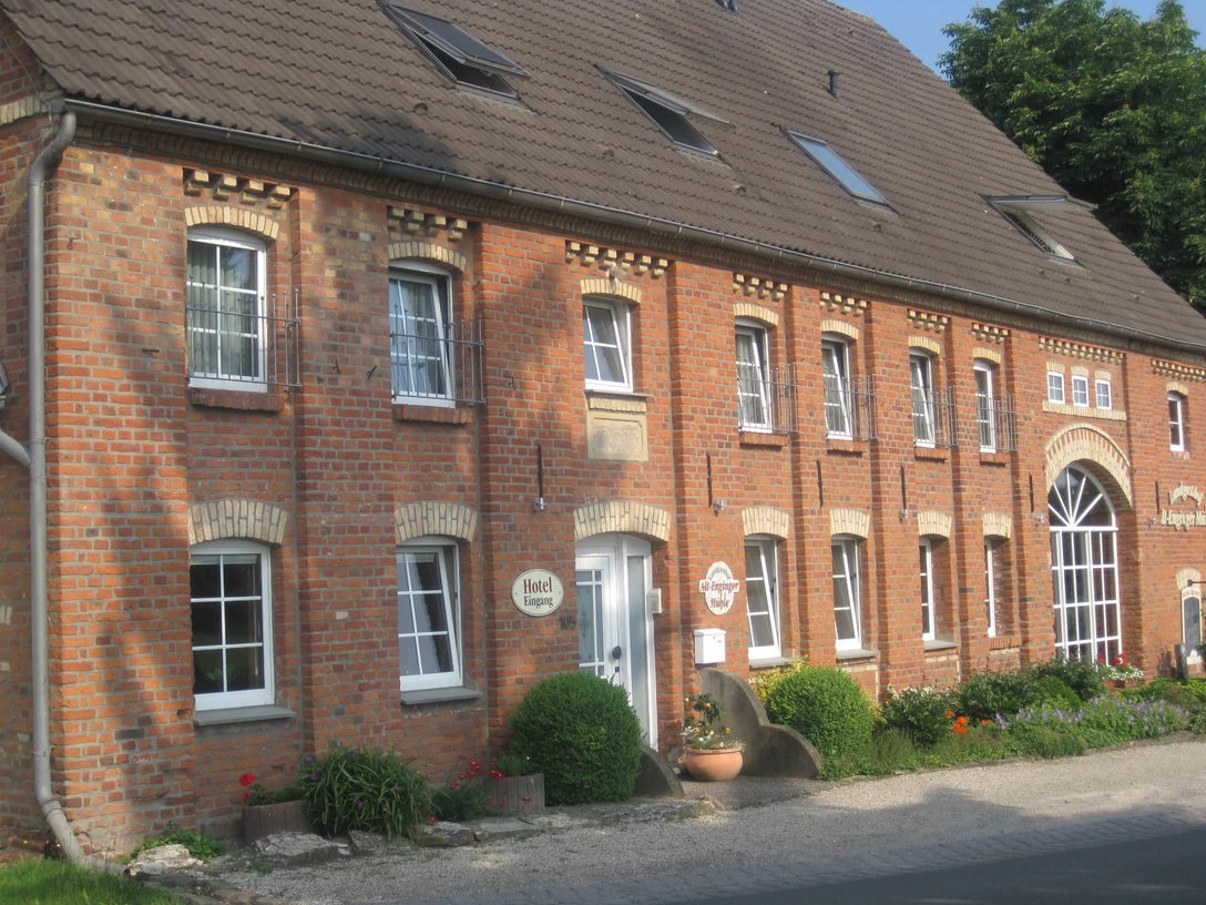 Alt-Enginger Mühle Backsteingebäude mit braunem Dach, weißen Fensterrahmen und gepflegtem Eingangsbereich, umgeben von Blumen.