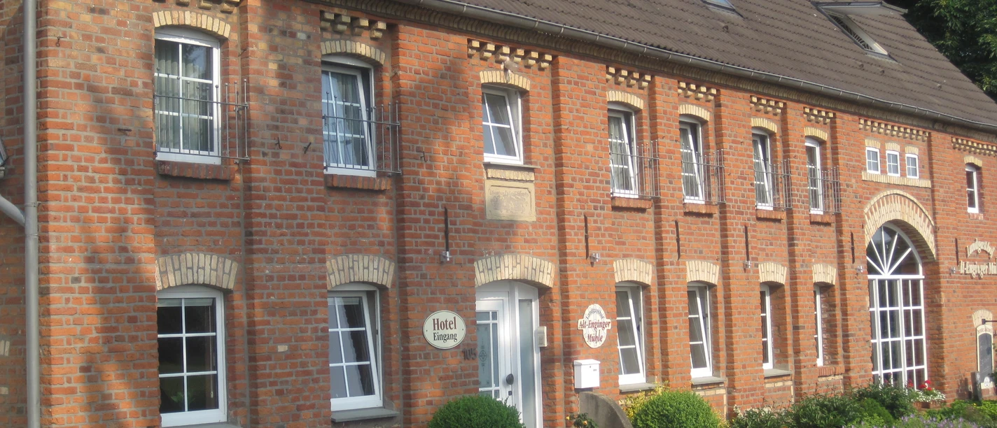 Alt-Enginger Mühle Backsteingebäude mit braunem Dach, weißen Fensterrahmen und gepflegtem Eingangsbereich, umgeben von Blumen.