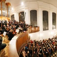 Blick auf einen mit Menschen gefüllten Kircheninnenraum, Empore und Orgel, vor der ein Chor und Musiker*innen stehen.View of a church interior filled with people, gallery and organ, in front of which a choir and musicians are standing.Vue d'un intérieur d'église rempli de gens, d'une tribune et d'un orgue devant lesquels se tiennent un chœur et des musiciens*.Gezicht op een kerkinterieur vol mensen, galerij en orgel, waarvoor een koor en muzikanten staan.
