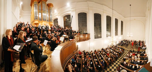 Blick auf einen mit Menschen gefüllten Kircheninnenraum, Empore und Orgel, vor der ein Chor und Musiker*innen stehen.