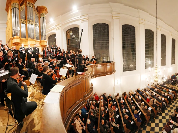 Blick auf einen mit Menschen gefüllten Kircheninnenraum, Empore und Orgel, vor der ein Chor und Musiker*innen stehen.