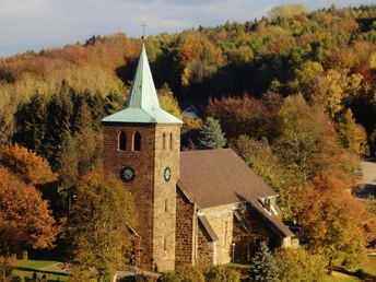 Steinernen Kirche mit grünem Turmdach, umgeben von buntem Herbstlaub und dichtem Waldhintergrund.