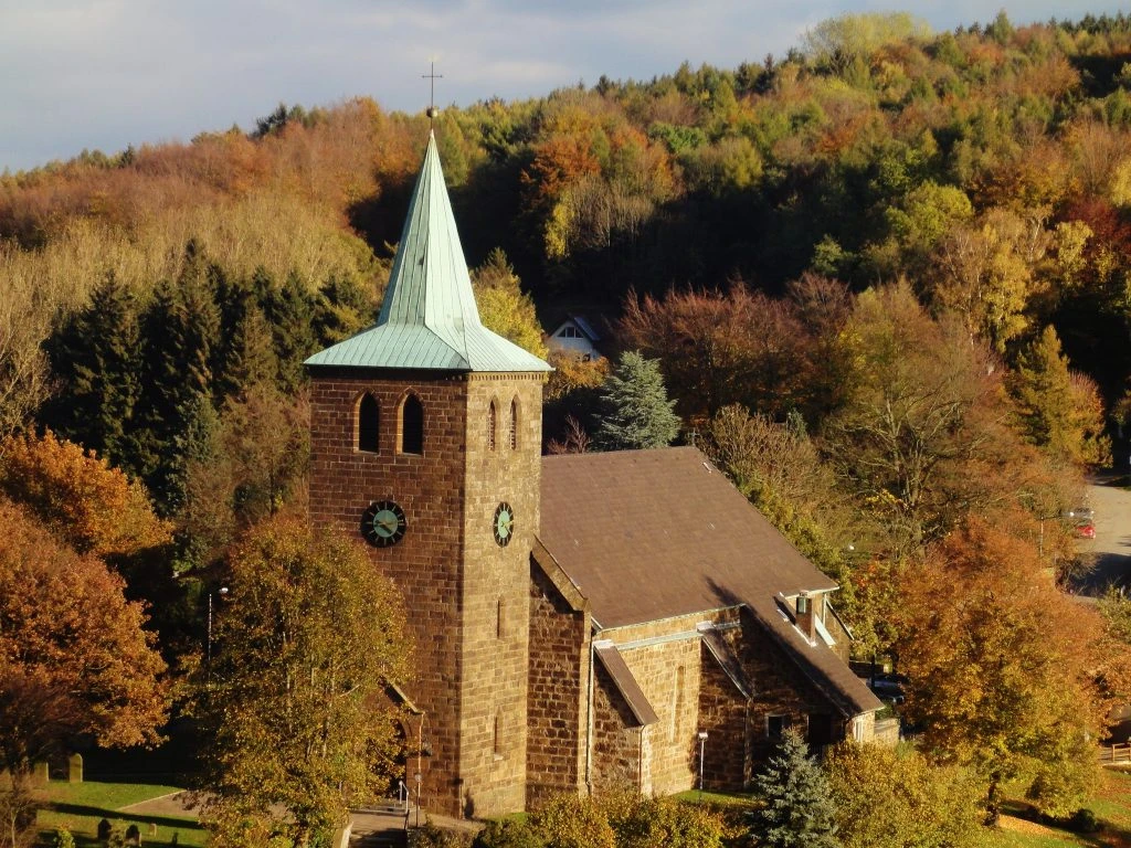 St.-Nikolaus-Kirche in Bergkirchen Steinernen Kirche mit grünem Turmdach, umgeben von buntem Herbstlaub und dichtem Waldhintergrund.