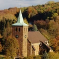 St.-Nikolaus-Kirche in Bergkirchen Steinernen Kirche mit grünem Turmdach, umgeben von buntem Herbstlaub und dichtem Waldhintergrund.