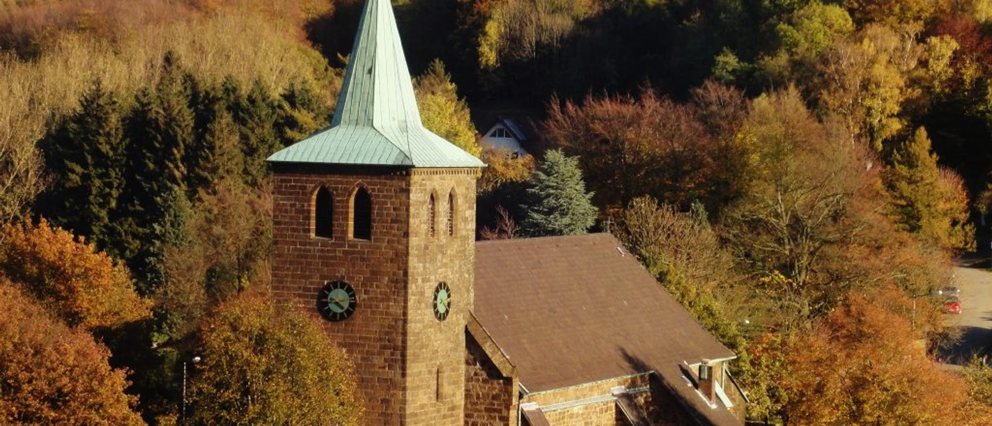 St.-Nikolaus-Kirche in Bergkirchen Steinernen Kirche mit grünem Turmdach, umgeben von buntem Herbstlaub und dichtem Waldhintergrund.