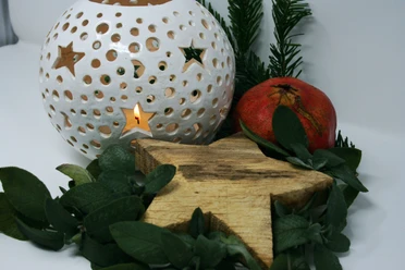 Eine weiße Laterne mit Sternenmustern leuchtet sanft neben einem roten Granatapfel und grünen Zweigen.