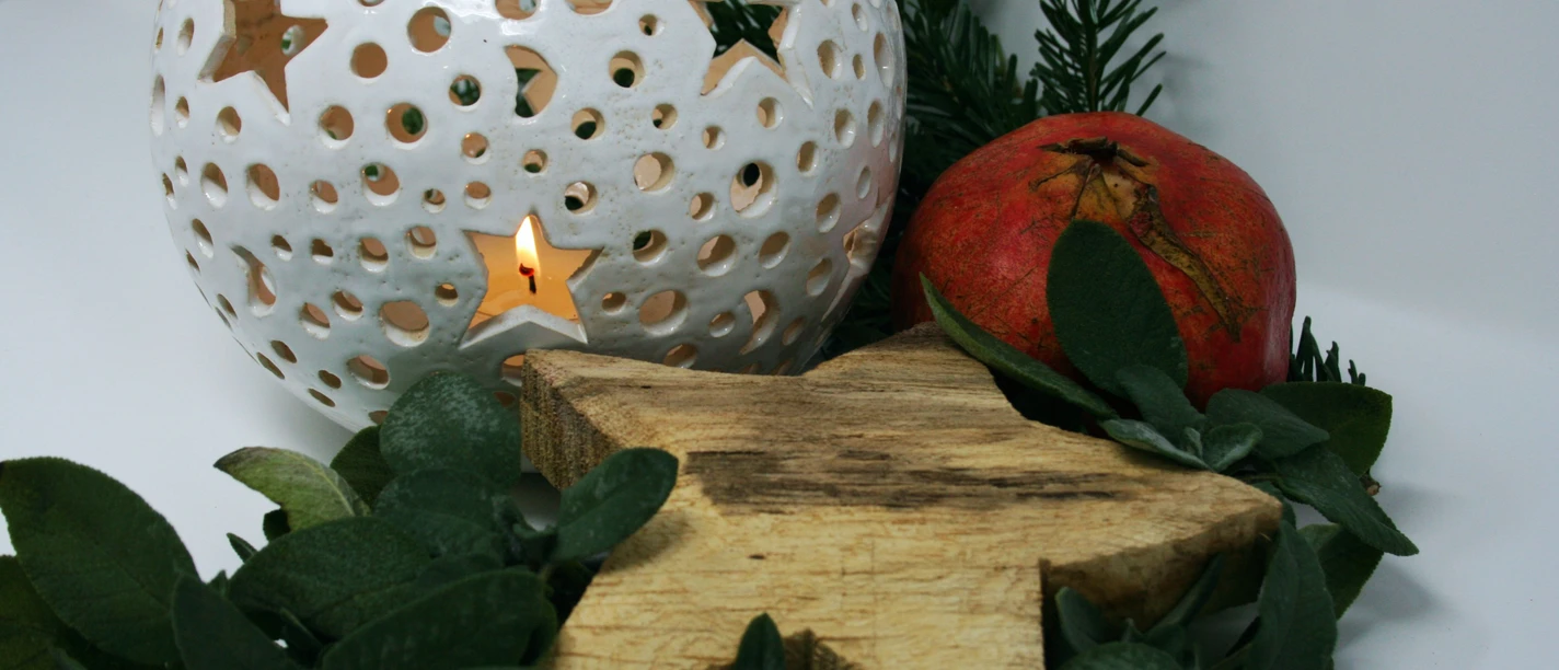Handkunstwerke-zur-Weihnachtszeit-web.jpg Eine weiße Laterne mit Sternenmustern leuchtet sanft neben einem roten Granatapfel und grünen Zweigen.
