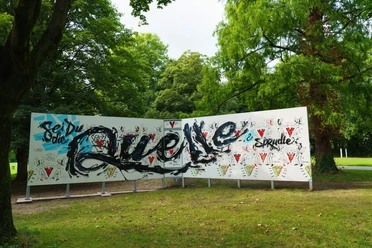 Graffitistern Paderborn Graffiti-Kunstwerk im Freien mit bunter Schrift und Mustern vor üppig-grünen Bäumen.