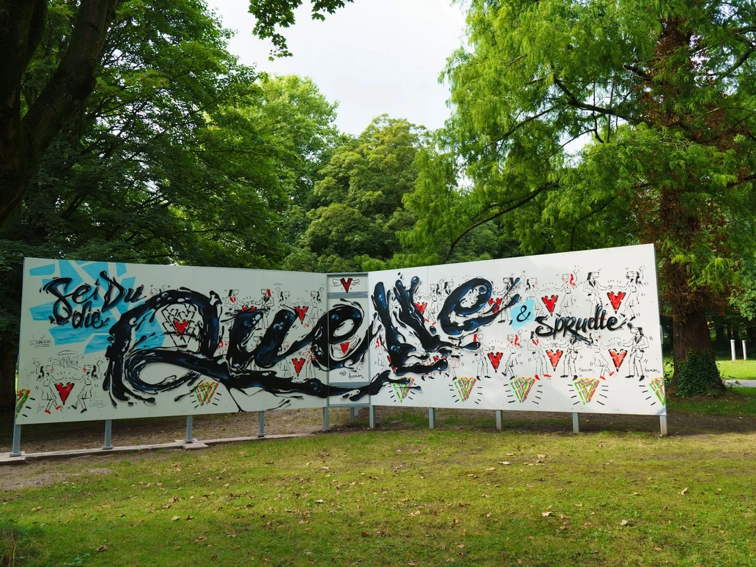Graffitistern Paderborn Graffiti-Kunstwerk im Freien mit bunter Schrift und Mustern vor üppig-grünen Bäumen.