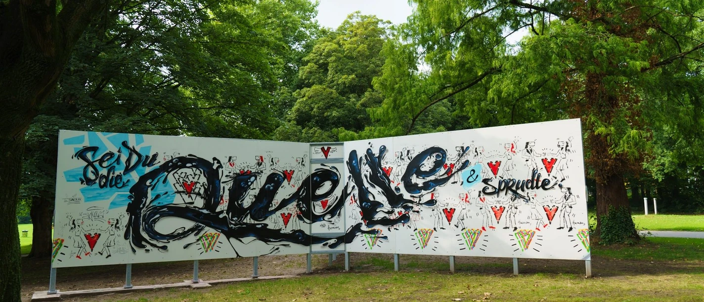 Graffitistern Paderborn Graffiti-Kunstwerk im Freien mit bunter Schrift und Mustern vor üppig-grünen Bäumen.