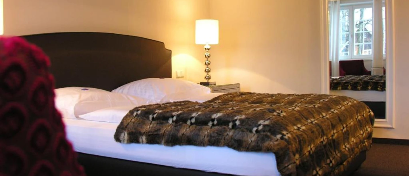 Doktorplatz.jpg Doppelzimmer mit stilvollem Design, weicher Tagesdecke, Nachttischlampe und gemütlicher Beleuchtung.