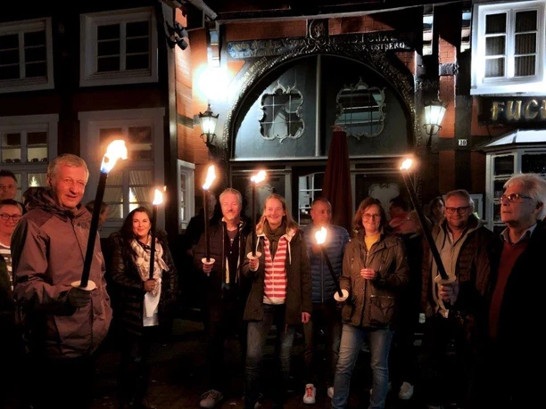 Fackel.jpg Menschen halten Fackeln vor einem beleuchteten, historischen Fachwerkgebäude in abendlicher Szenerie.
