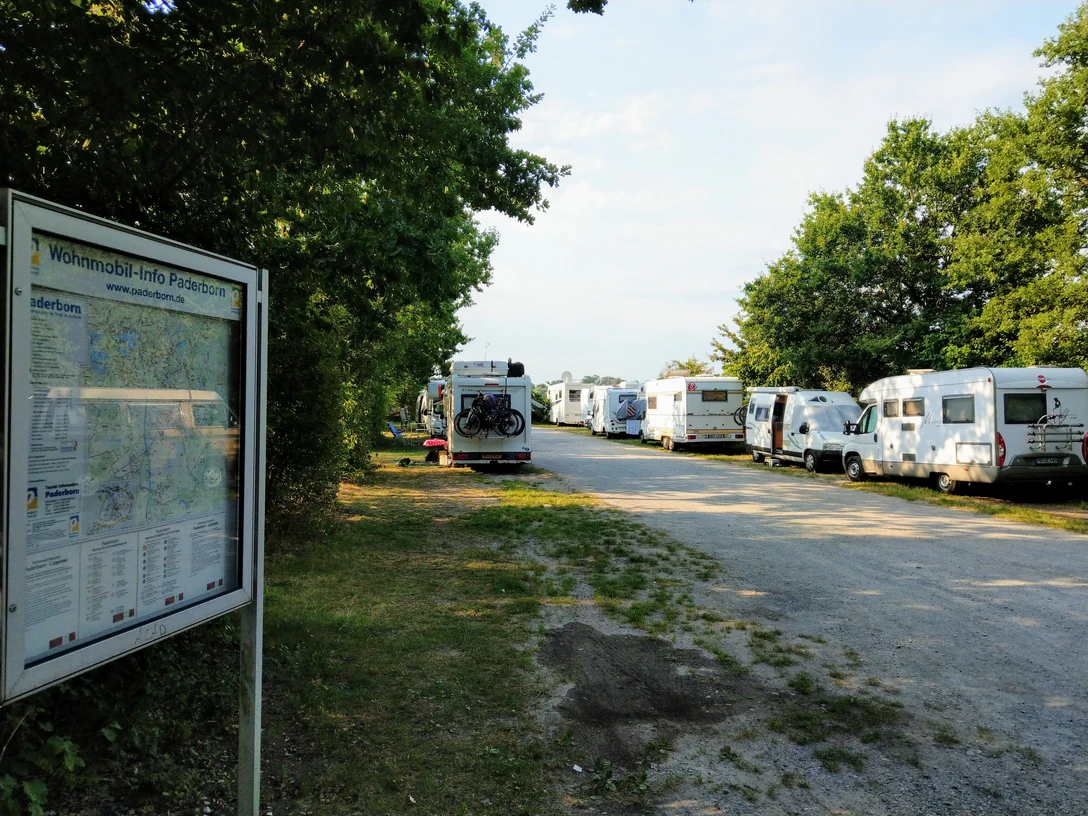 Reihen von Wohnmobilen parken entlang eines schattigen Weges, umgeben von Bäumen und grüner Natur.