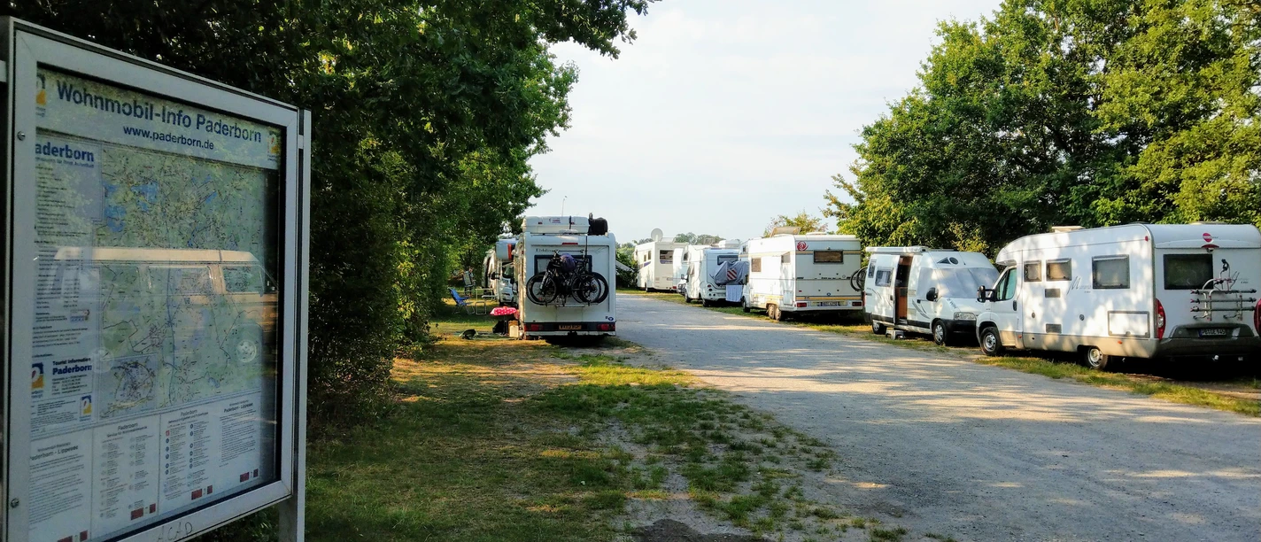 Reihen von Wohnmobilen parken entlang eines schattigen Weges, umgeben von Bäumen und grüner Natur.
