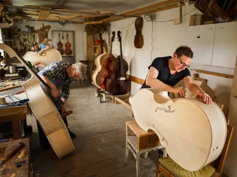 Erlebniswelt Musikinstrumentenbau Zwei Männer arbeiten konzentriert an großen, unlackierten Musikinstrumenten in einer Holzwerkstatt.Two men work intently on large, unpainted musical instruments in a wood workshop.Dva muži v dřevařské dílně soustředěně pracují na velkých nelakovaných hudebních nástrojích.Dwóch mężczyzn intensywnie pracuje nad dużymi, niepomalowanymi instrumentami muzycznymi w warsztacie drzewnym.Twee mannen werken aandachtig aan grote, onbeschilderde muziekinstrumenten in een houtwerkplaats.Due uomini lavorano intensamente su grandi strumenti musicali non verniciati in un laboratorio di legno.