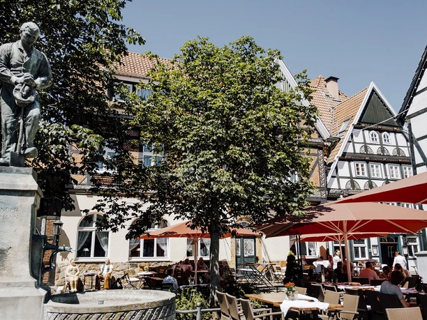 Statue neben einem Brunnen, umgeben von gemütlichen Café-Terrassen mit roten Sonnenschirmen im Sommersonnenschein.
