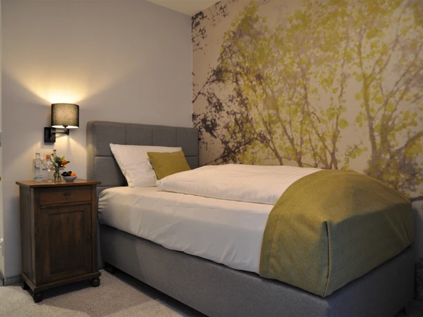 Ein gemütliches Einzelzimmer mit modernem grauem Bett, Holznachttisch und Fototapete eines Baumes.