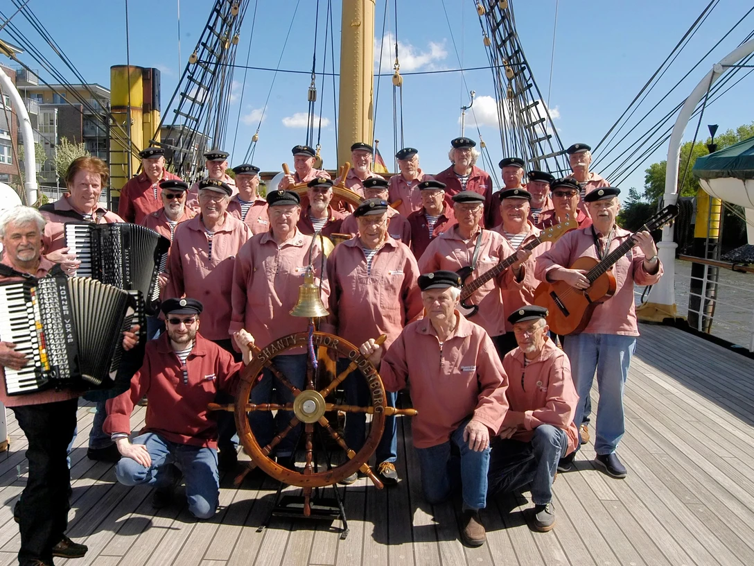 shanty-chor-oldenburg.jpg Shanty-Chor in roten Hemden und Matrosenmützen auf einem Segelschiff, mit Akkordeon und Gitarren.
