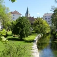 Flusslauf im grünen Park, umgeben von Bäumen und historischen Gebäuden mit Kirchturm im Hintergrund.