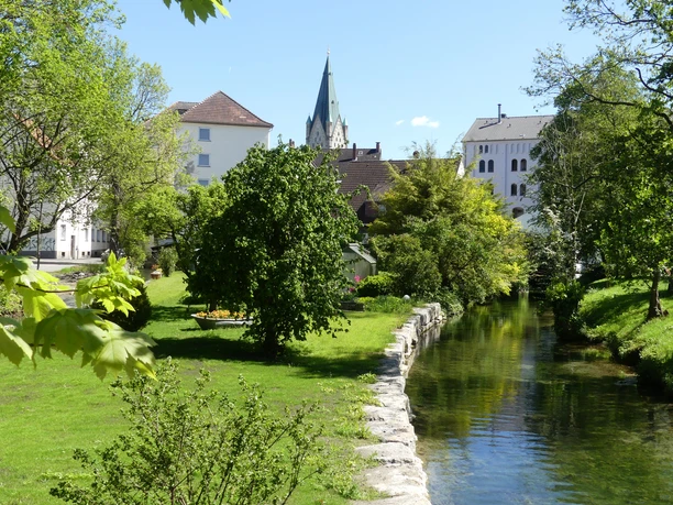 Flusslauf im grünen Park, umgeben von Bäumen und historischen Gebäuden mit Kirchturm im Hintergrund.