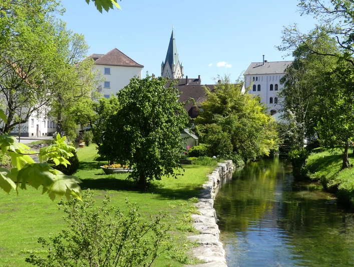 Flusslauf im grünen Park, umgeben von Bäumen und historischen Gebäuden mit Kirchturm im Hintergrund.