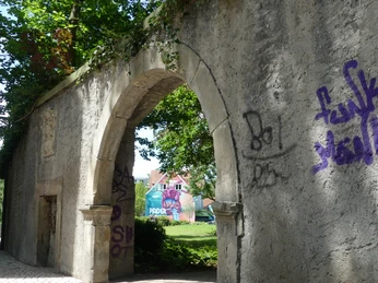 Altes Steinportal mit Graffiti, überwuchert von Pflanzen. Durchblick zum bunt bemalten Gebäude im Garten.
