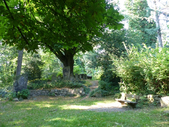 Natursteingrotte mit großen Bäumen, Sitzbank und umgeben von üppigem Grün in einem kleinen Park.