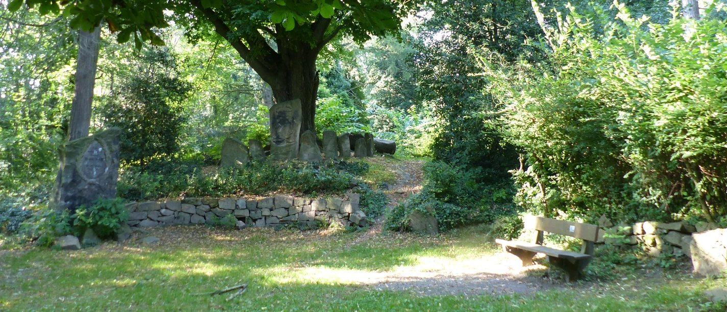 Steingrotte©Margarete Wißmann.JPG Natursteingrotte mit großen Bäumen, Sitzbank und umgeben von üppigem Grün in einem kleinen Park.