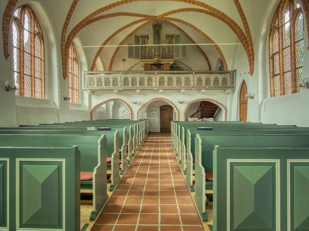 Blick durch das Kirchenschiff Blick durch das Kirchenschiff mit grünen Holzbänken zur hölzernen Orgelempore unter dem Gewölbe.