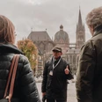 Mann mit Kappe steht auf dem Katschhof vor dem Aachener Dom und erzählt einer Gruppe von Personen etwas.