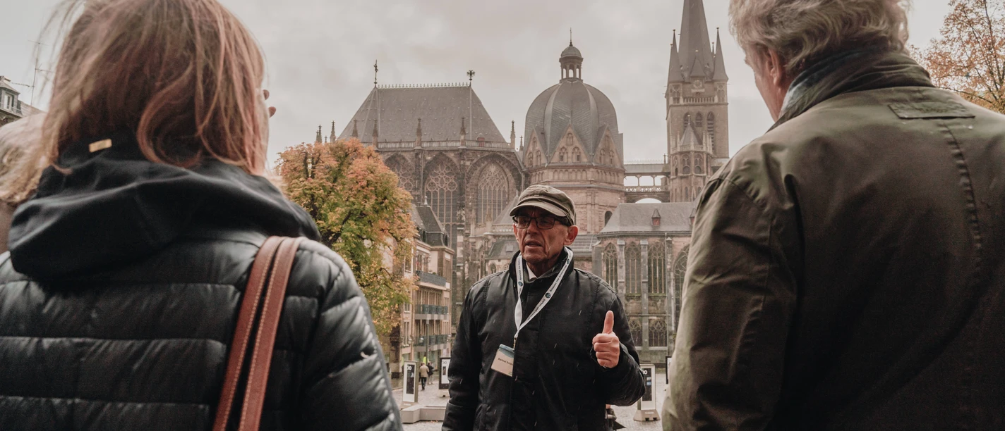 Mann mit Kappe steht auf dem Katschhof vor dem Aachener Dom und erzählt einer Gruppe von Personen etwas.