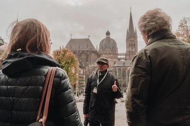 2021-10 Stadtfuehrung (c) ats_tom tietz (10).jpg Mann mit Kappe steht auf dem Katschhof vor dem Aachener Dom und erzählt einer Gruppe von Personen etwas.
