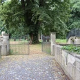 Historisches Tor mit schmiedeeisernen Flügeltüren führt in eine von Bäumen gesäumte, ruhige Gartenanlage.