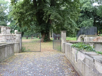 Historisches Tor mit schmiedeeisernen Flügeltüren führt in eine von Bäumen gesäumte, ruhige Gartenanlage.