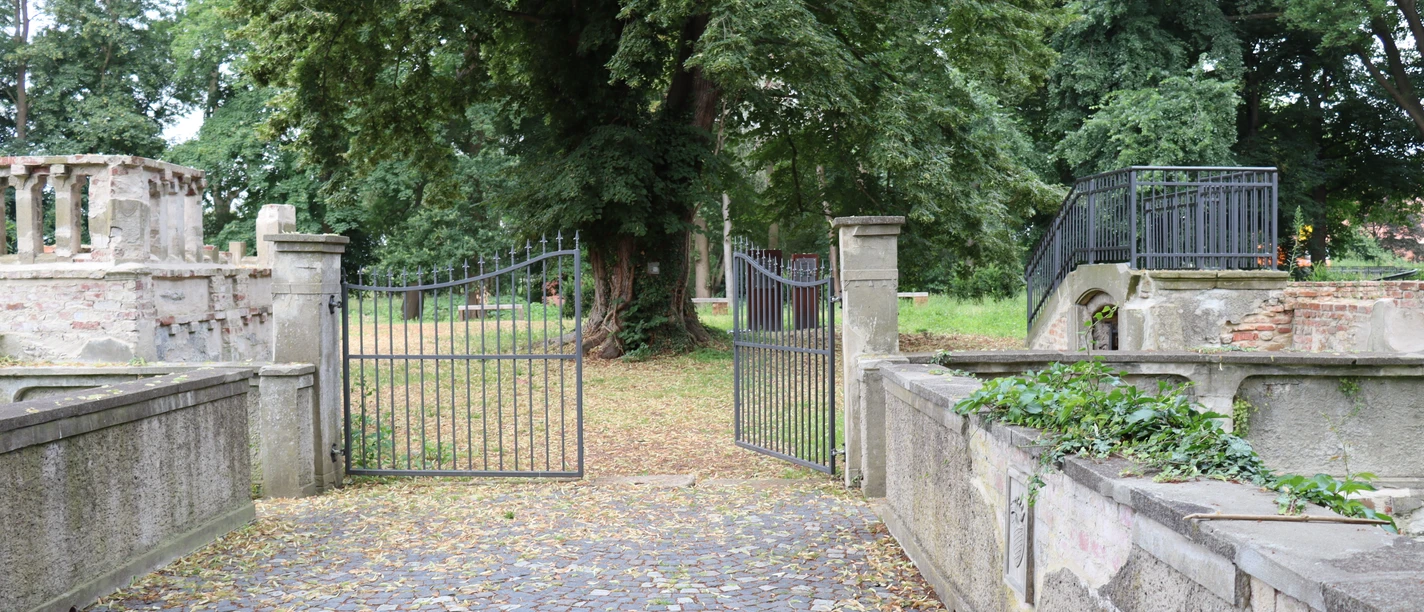 Ellerburg Historisches Tor mit schmiedeeisernen Flügeltüren führt in eine von Bäumen gesäumte, ruhige Gartenanlage.