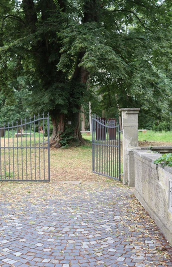 Ellerburg Historisches Tor mit schmiedeeisernen FlĂ¼geltĂ¼ren fĂ¼hrt in eine von Bäumen gesäumte, ruhige Gartenanlage.