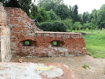 Ziegelruinen einer mittelalterlichen Burgmauer mit zwei Fensteröffnungen, umgeben von grüner Landschaft.