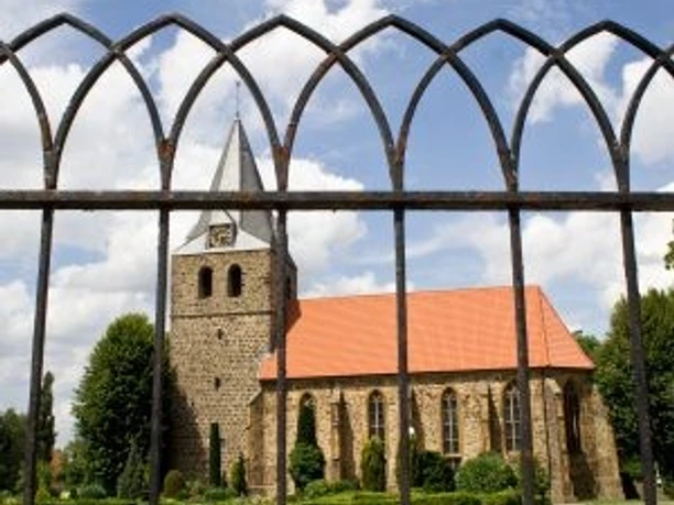 Gehlenbeck-Kirche.jpg Eine historische Kirche aus Stein mit rotem Dach hinter einem schwarzen, schmiedeeisernen Gitter.