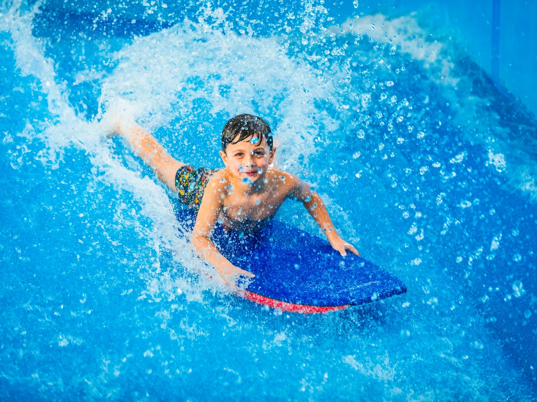 Ein Kind gleitet lachend auf einem blauen Bodyboard über eine Wasseroberfläche.A child glides across the water surface on a blue bodyboard, laughing.