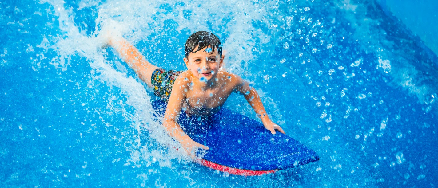 Ein Kind gleitet lachend auf einem blauen Bodyboard über eine Wasseroberfläche.A child glides across the water surface on a blue bodyboard, laughing.