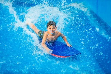 FlowRider Ein Kind gleitet lachend auf einem blauen Bodyboard über eine Wasseroberfläche.