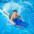FlowRider Ein Kind gleitet lachend auf einem blauen Bodyboard über eine Wasseroberfläche.