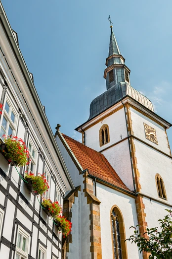 Rietberg-St-Joh-Baptist-K-prowiGT-Wallenfang.jpg Dreistöckiges Fachwerkgebäude mit Blumenkästen, daneben imposante weiße Kirche mit spitzem Turm.