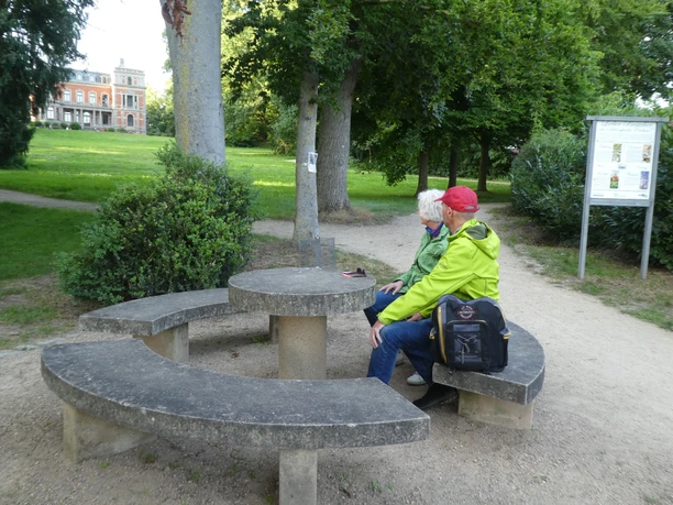 Rastplatz Schloss Etelsen Zwei Personen sitzen auf einer betonierten Sitzgruppe in einem grünen Park mit historischen Gebäuden.