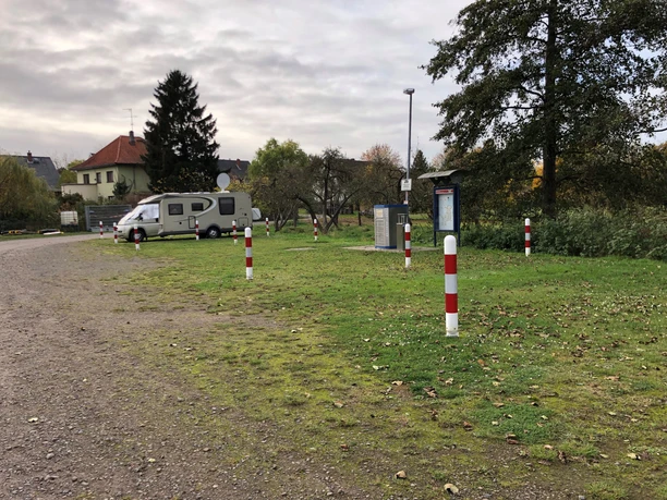 Wohnmobilstellplatz mit gepflastertem Untergrund, umgeben von Bäumen und Wohnhäusern im Hintergrund.