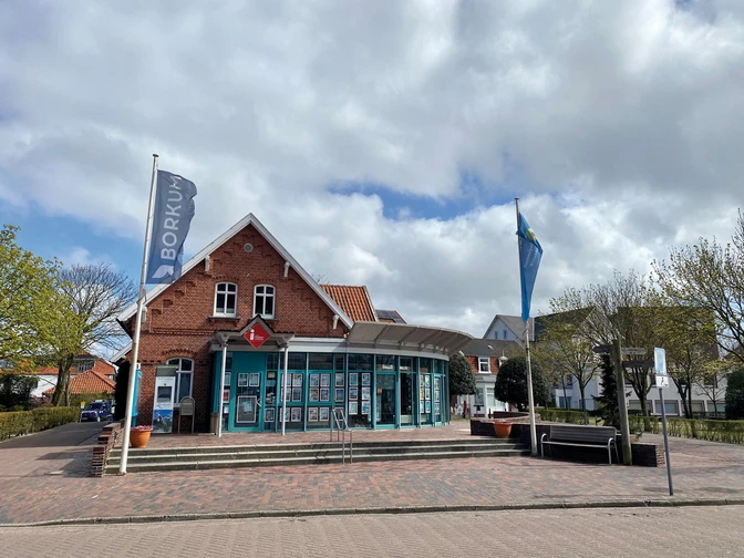 Das Tourist-Informationszentrum im Backsteingebäude mit blauer Fassade und Flaggen davor. Der Himmel ist bewölkt.The tourist information center in the brick building with a blue façade and flags in front of it. The sky is cloudy.