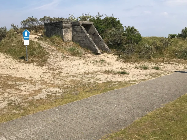 Alter Bunker Ein alter Bunker aus Beton, umgeben von Sand und Gras, erhebt sich unter klarem Himmel am Wegesrand.