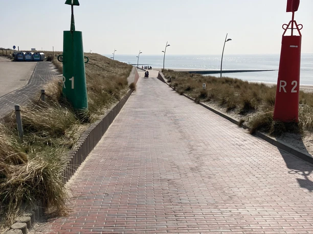 Zugang Nordstrand Ein asphaltierter Weg führt durch grasbewachsene Dünen hinab zum Meer, flankiert von grüner und roter Boje.