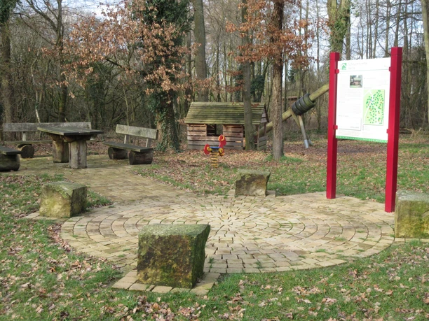 Scheunenviertel Wellie Spielplatz im Grünen mit Holztischen und Bänken, Schaukel und Infotafel, umgeben von Bäumen.